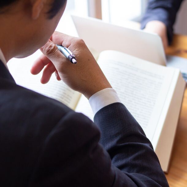 Person holding a pen looking down at a book, another person sitting across typing on a laptop