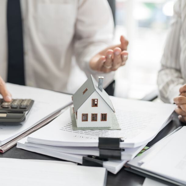 Person typing on a calculator, small house diorama sitting on top of a stack of papers on a desk