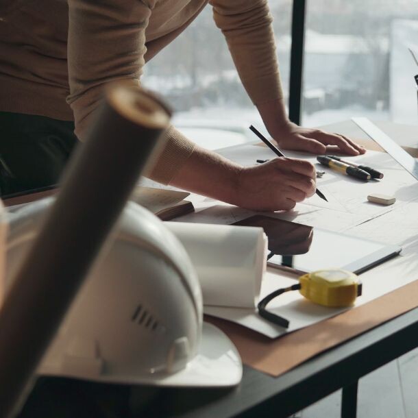 Person writing on a piece of paper on a desk with plants, posters, and a hard hat