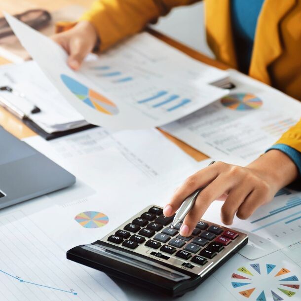 Person sitting behind a desk facing reports, a clipboard, a laptop and a calculator. On their right hand, they are holding a sheet of paper, and on the left, they are typing on the large calculator.