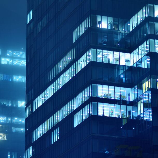 In a somewhat-dark night sits a blue tall builing and shines through a portion of the windows.