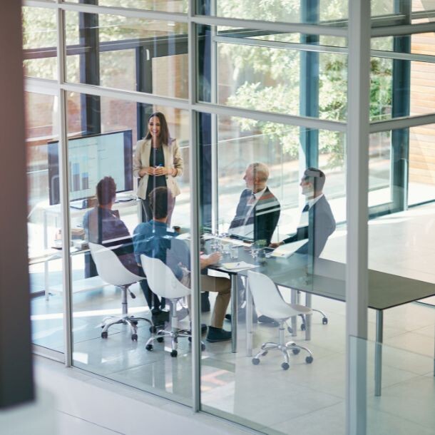 Behind the glass doors of an office is a group of five people discussing by a large white meeting room, all facing a presentation screen.