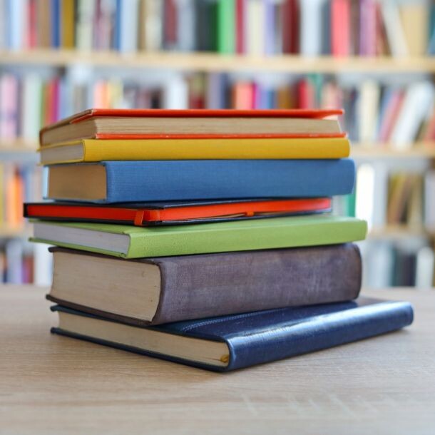 Pile of solid-colored books sitting at a table in front of a libary shelf.