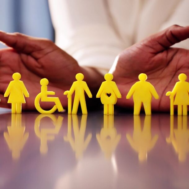 Diversely-shaped human icons stand in yellow on top of a partially reflective glass table. A person stands with their hands behind them in an embracing format.