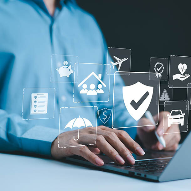 A person typing on their laptop's keyboard, wearing a blue dress shirt. On top of the laptop are white icons; the largest ones include a shield, a hand holding a heart, a list, a home with people insise, an umbrella, a plane, a car and a briefcase.