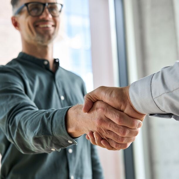 A hand coming from the right side of the screen comes in a white dress shirt to participate in a handshake with a man, who is wearing classes, a darker dress shirt and smiling.