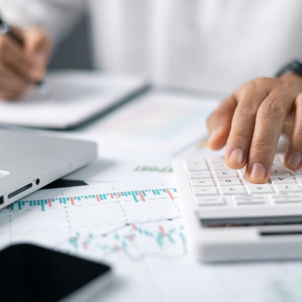 A person dressed in a white dress shirt, wearing a watch, holding apen on their right hand (scribbling down notes) and they type on a white calculator with their left hand. There are also a laptop, a cellphone and a sheet with graphs on top of their desk.