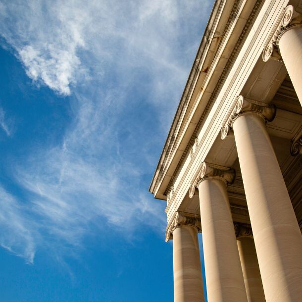 Shot from the ground up, half of the frame includes a tall building made up of stone columns, and the other half is a shot of blue skies covered by a touch of clouds.