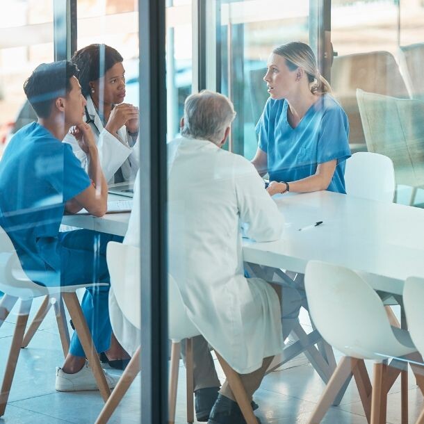 Behind the glass doors of an office is a group of four people discussing by a large white meeting room.