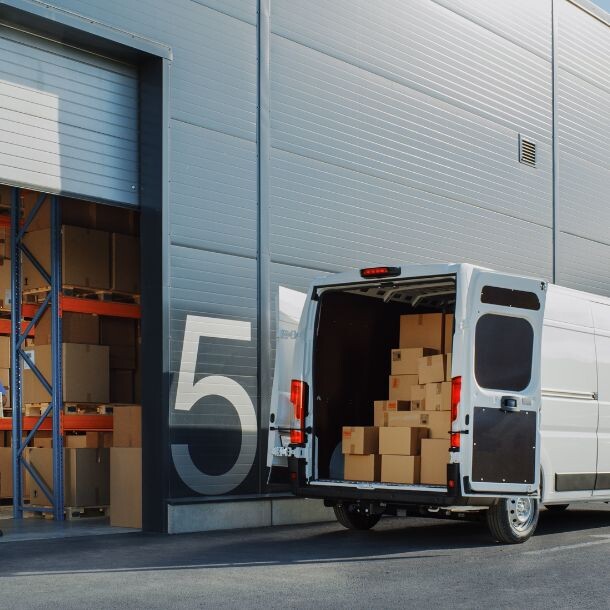 In front of a warehouse full with boxes is a white van getting loaded by two employees wearing blue.