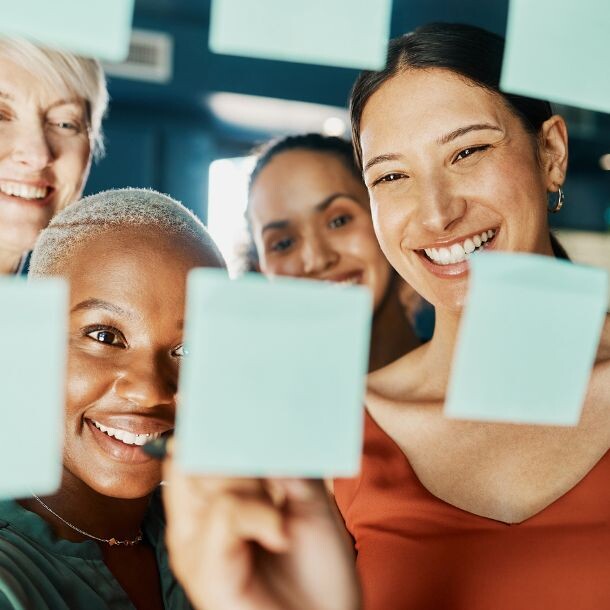 A diverse group of people smile at sticky notes, which stand in front of them and right at the forefront og the image.