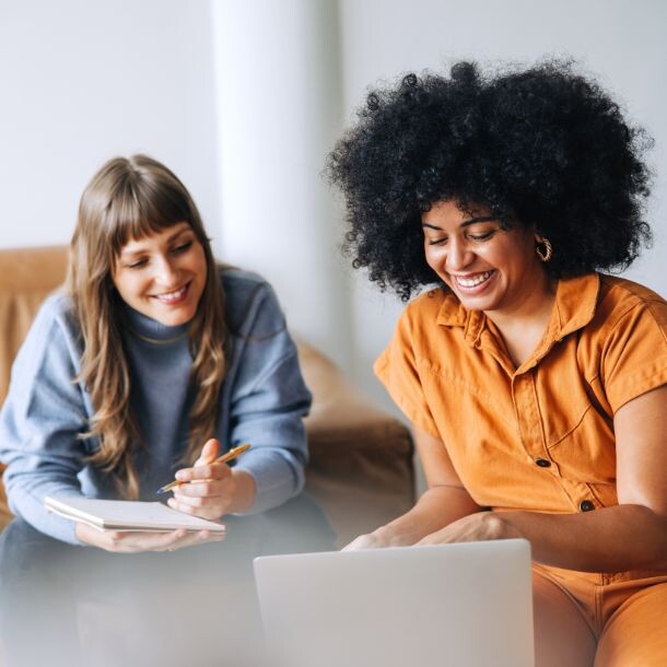Two women in a casual setting sitting side to side looking at the same laptop screen.