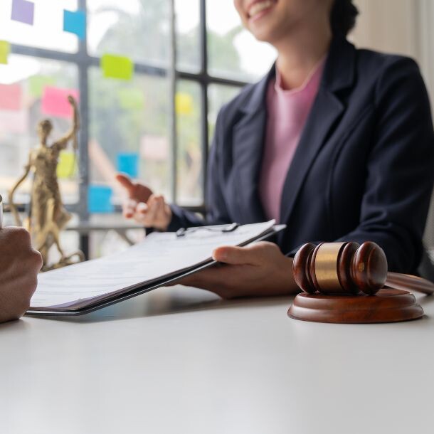 A person signing a contract that is being held by a woman sitting at a desk. To the woman's left side is a court hammer.