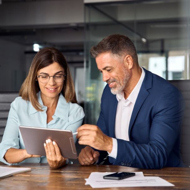 A woman showing a man (both dressed professionally) something on a tablet. Both sit side to side at a desk, facing documents and the man's cellphone.