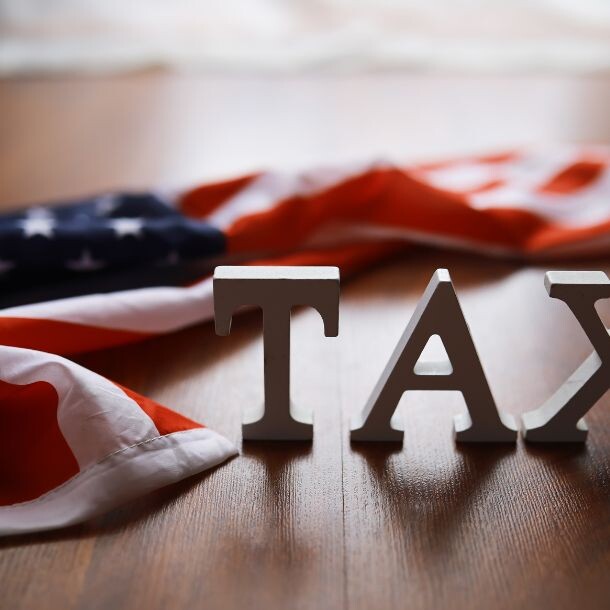 A wooden table where sit the word "TAX" made of wooden pieces, and a slightly-crumpled American flag.