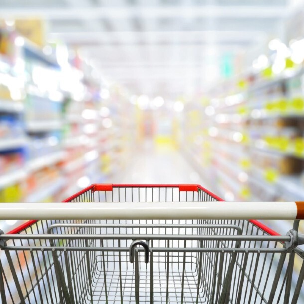 Empty shopping cart in a supermarket aisle with blurred shelves of consumer goods.